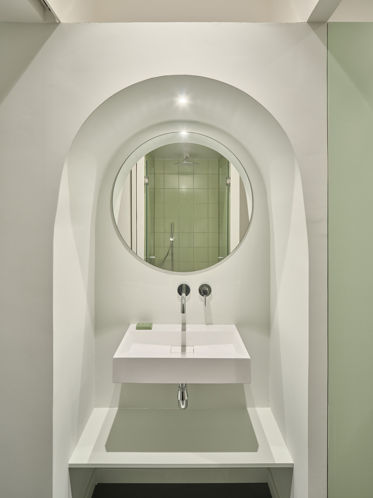 Arched niche in the bathroom of CRÜ’s “La Dolors” renovation, with a minimalist wall-mounted sink and a circular mirror reflecting the green-tiled shower